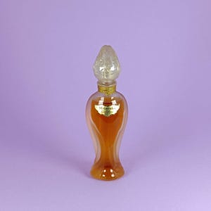 Op de afbeelding: Een vintage glazen flesje Mitsouko parfum met een gouden etiket en een doorzichtige glazen dop in de vorm van een bloemknop. De fles is gevuld met een licht amberkleurige vloeistof.