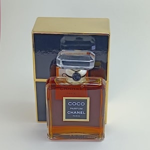 Puede incluir: Un frasco de perfume Coco Chanel Parfum de vidrio transparente con una tapa dorada y una etiqueta dorada. El frasco está sobre una caja dorada y azul.