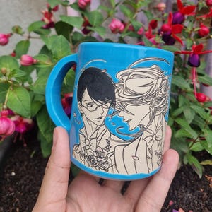Könnte beinhalten: Eine türkisfarbene Keramiktasse mit einer Schwarz-Weiß-Illustration von zwei Figuren im Anime-Stil. Die Tasse hat einen gebogenen Henkel. Der Hintergrund zeigt grünes Laub und rosa und rote Blüten.