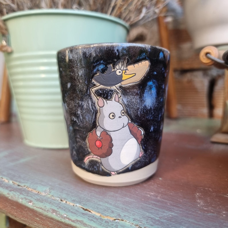 Może przedstawiać: Ceramiczny kubek z czarno-niebieską, nakrapianą glazurą. Kubek przedstawia kresk&oacute;wkową ilustrację szarej myszy trzymającej rękawicę do baseballu i czarnego ptaka z ż&oacute;łtym dziobem. Kubek stoi na drewnianej powierzchni.