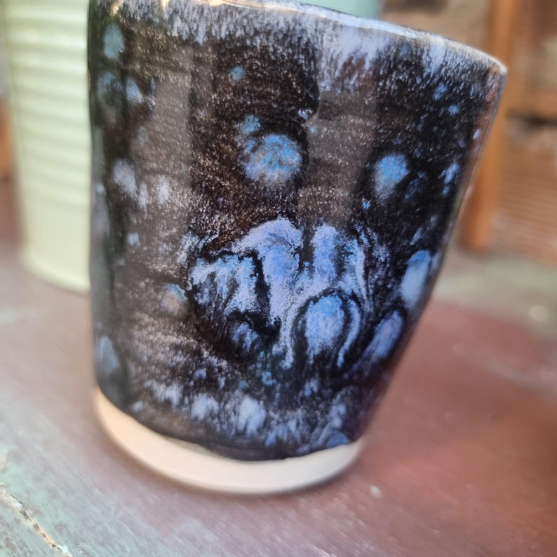 Może przedstawiać: Ręcznie wykonany ceramiczny kubek z unikalnym szkliwem. Kubek ma ciemny, prawie czarny wygląd zewnętrzny z niebieskimi i białymi akcentami. Podstawa jest czysto biała, a kubek ma lekko nieregularny kształt, co sugeruje ręczne wykonanie.