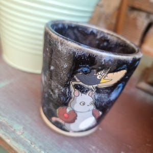 Może przedstawiać: Ceramiczny kubek z ciemnym, nakrapianym zewnętrzem i jasną podstawą. Kubek przedstawia kresk&oacute;wkową ilustrację szarego stworzenia z rękawicą do baseballu i czarnego ptaka z ż&oacute;łtym dziobem.