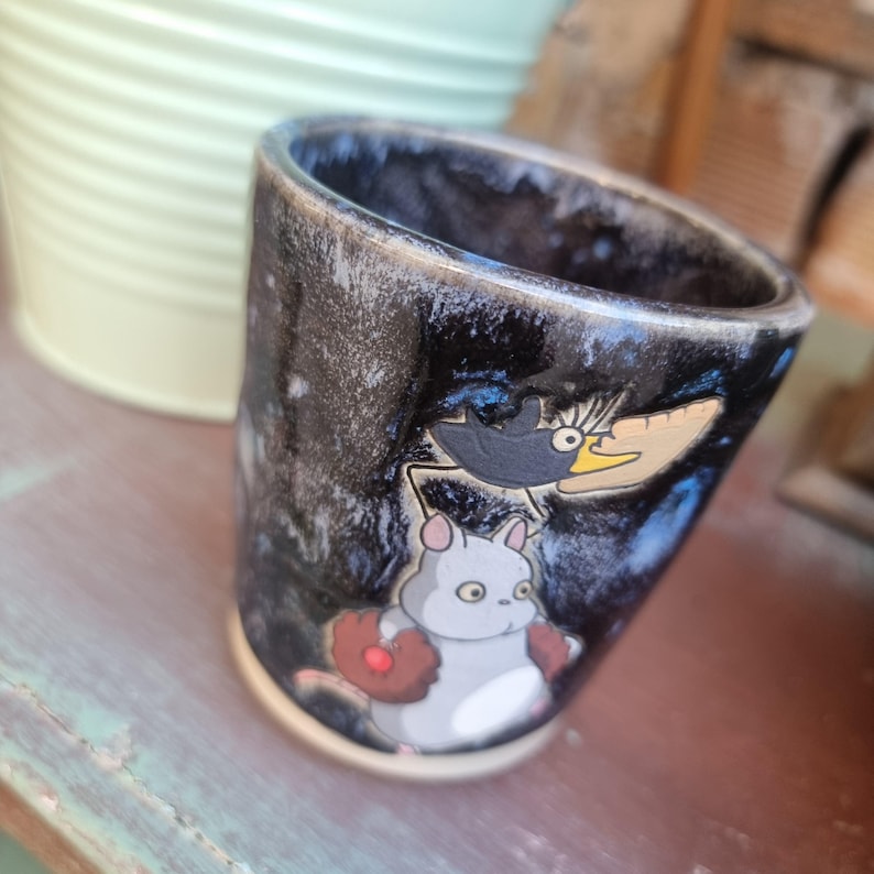 Może przedstawiać: Ceramiczny kubek z ciemnym, nakrapianym zewnętrzem i jasną podstawą. Kubek przedstawia kresk&oacute;wkową ilustrację szarego stworzenia z rękawicą do baseballu i czarnego ptaka z ż&oacute;łtym dziobem.