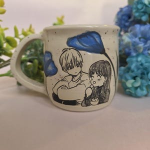 Può includere: Una tazza in ceramica con finitura bianca maculata, con un'illustrazione disegnata a mano di due personaggi in stile anime e accenti floreali blu. La tazza ha un manico curvo e una base arrotondata.