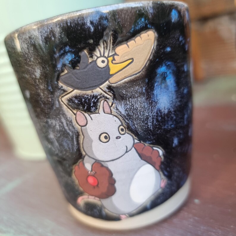 Może przedstawiać: Ceramiczny kubek z czarno-niebieską, nakrapianą glazurą. Kubek przedstawia kresk&oacute;wkową ilustrację czarnego ptaka i białego stworzenia z brązowymi rękawicami. Kubek ma kremową podstawę.