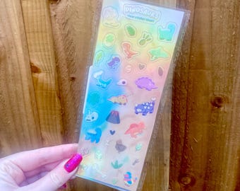 May include: A clear sticker sheet with colorful dinosaur illustrations. The sheet includes various dinosaur designs, a volcano, and other related graphics. The text "DINOSAURS clear sticker sheet" is printed at the top. The stickers are held in a clear plastic sleeve.