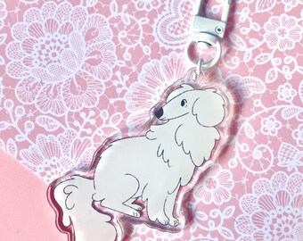 May include: A clear acrylic keychain featuring a white cartoon dog with black accents. The dog has fluffy fur and is attached to a silver-colored clasp. The background is a pink lace pattern.