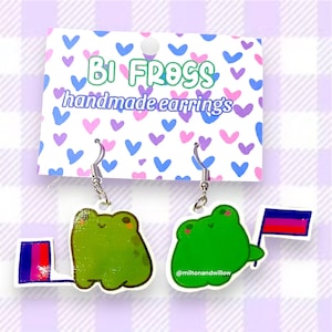 May include: A pair of handmade earrings featuring green frog designs. One frog holds a flag with the bisexual pride colors. The earrings hang from silver-colored hooks. The backing card reads "Bi Frogs handmade earrings."