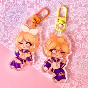 May include: Two anime-style acrylic keyrings featuring characters with orange hair and sailor-style outfits in purple and white. Each keyring has a different pose and is attached to a coloured clasp, one orange and one yellow.