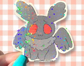 May include: A glittery sticker of a cartoon moth with large red eyes and wings. The moth is grey with iridescent glitter accents. The sticker has a white border and is set against a pink and white gingham background. The text "@miltonandwillow" is at the bottom.