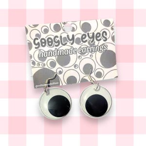 Può includere: Un paio di orecchini con occhietti a palla con ganci argentati. Gli orecchini sono fatti a mano e presentano uno sfondo bianco con un cerchio nero al centro. Gli orecchini sono confezionati su una carta con il testo "googly eyes handmade earrings" e l'account social "@miltosandwillow".