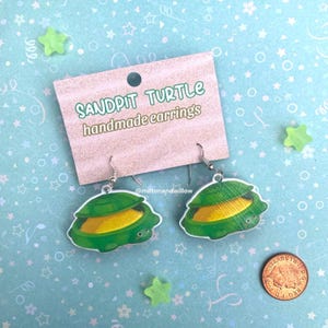 May include: A pair of handmade earrings featuring a sandpit turtle design. The earrings are made of green and yellow acrylic and have silver hooks. The earrings are displayed on a card that says "Sandpit Turtle handmade earrings" and includes the social media handle @miltonandwillow.