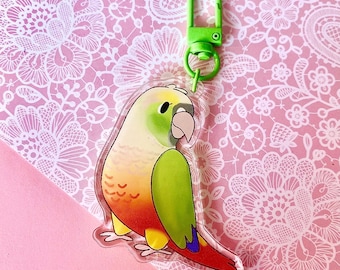 May include: A colorful parrot-shaped keychain with a green clip. The parrot has a white beak, green and yellow head, green and orange wings, and a red tail. The background is a pink surface with a white floral pattern.