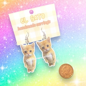 Op de afbeelding: Een paar oorhangers met een schattig oranje tabby-kittens met een witte borst. De oorhangers zijn gemaakt van helder acryl en hebben zilveren haakjes. De oorhangers zijn gelabeld met "El Gato handmade earrings" en "@miltonandwillow".