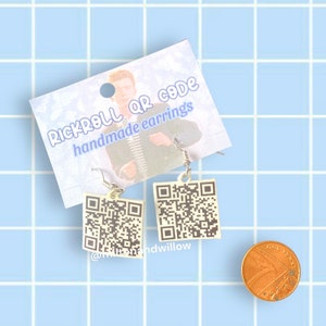 May include: A pair of handmade earrings featuring a white square with a black QR code printed on it. The earrings are hanging from silver hooks. The earrings are displayed on a white card with the text "Rickroll QR code handmade earrings" and a picture of a man.