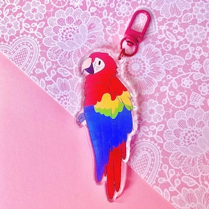 May include: A colorful parrot keychain with a red, yellow, blue, and green design. The keychain has a pink clasp and is made of clear acrylic. The parrot is a cartoon illustration.