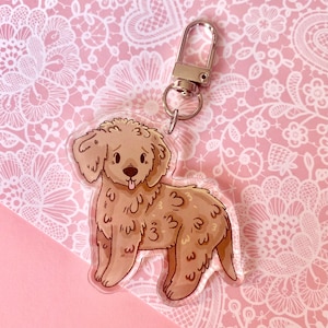 May include: A clear acrylic keychain featuring a cartoon illustration of a light brown dog with the number 3 printed on its fur. The keychain has a silver-colored clasp for attaching to keys or bags. The background is a pink lace pattern.