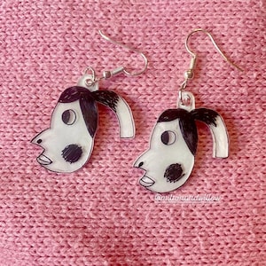 May include: A pair of white earrings featuring black cartoon faces with black hair. The earrings are shaped like a person's head with a curved line extending from the side of the head.