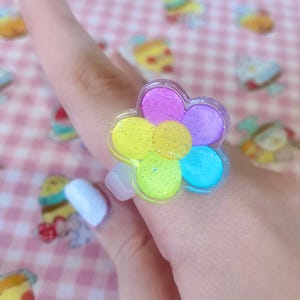 May include: A flower-shaped ring with glitter detailing. The ring has petals in yellow, green, blue, and purple. The ring is made of clear plastic and is worn on a finger.