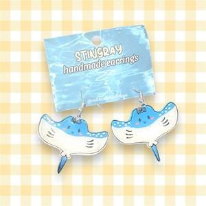 May include: A pair of handmade stingray earrings with blue and white paint and silver hooks. The earrings are on a yellow and white checkered background. The text on the card reads "STINGRAY handmade earrings @miltonandwillow"