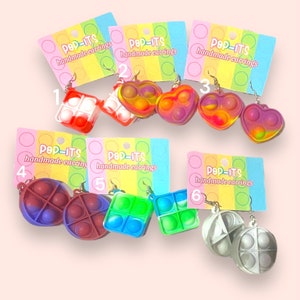 Könnte beinhalten: Sechs Paar handgefertigte Pop-It-Ohrringe. Die Ohrringe sind aus Silikon und in verschiedenen Farben und Formen erhältlich, darunter quadratisch, herzförmig und rund. Die Ohrringe sind mit den Zahlen 1 bis 6 gekennzeichnet.