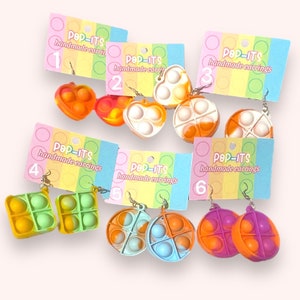 May include: Six pairs of handmade pop-it earrings. The earrings are made of silicone and come in a variety of colors and shapes, including heart, circle, and square. The earrings are labeled with numbers 1 through 6.