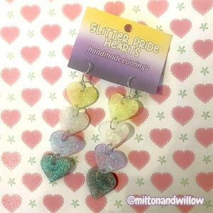 Puede incluir: Pendientes hechos a mano Glitter Pride Hearts. Estos pendientes colgantes presentan dijes en forma de corazón en un degradado de colores, incluyendo amarillo, blanco, lavanda y verde azulado. Los pendientes están suspendidos de ganchos plateados, con un fondo estampado.