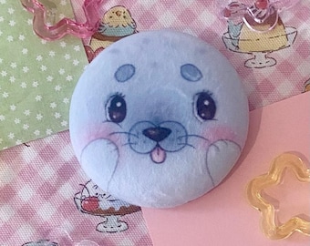Puede incluir: Un juguete de peluche redondo con forma de foca, de color azul claro, con ojos grandes y mejillas rosadas. La foca tiene una pequeña lengua que sobresale. El fondo incluye tela estampada rosa y verde y decoraciones de plástico en forma de estrella.