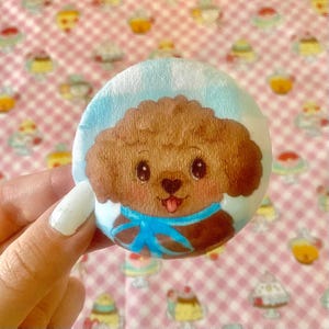 Puede incluir: Un botón redondo, celeste y marrón con un caniche de dibujos animados con una bufanda azul. El caniche tiene una expresión feliz con la lengua rosa fuera. El fondo es un patrón de cuadros rosas y blancos con ilustraciones de cupcakes.
