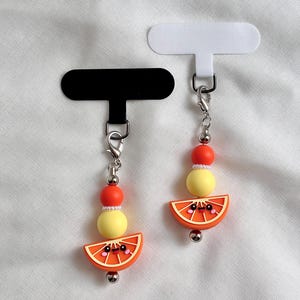 Könnte beinhalten: Zwei Anhänger mit einer lächelnden Orangenscheibe, einer gelben Perle, einer orangefarbenen Perle und einer kleinen weißen Zwischenperle. Jeder Anhänger hängt an einem silberfarbenen Karabinerverschluss an einer T-förmigen Karte, schwarz und weiß.