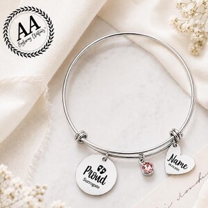 May include: A silver-toned expandable bangle bracelet featuring three charms: a round disc engraved with the text Proud Surrogate and baby footprints, a small pink crystal charm, and a heart-shaped charm engraved with Name and 06.23.2025.
