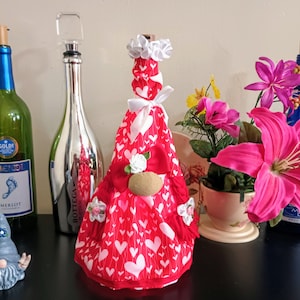 May include: A collection of decorative wine bottles and accessories. A red bottle cover with white hearts and a gnome-like design is featured. Other bottles are green and blue, with a silver bottle stopper. Pink flowers and a small gnome figurine are also present.