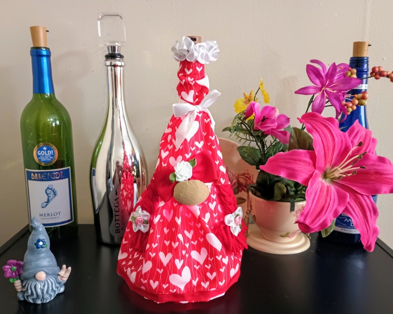 May include: A collection of decorative wine bottles and accessories. A red bottle cover with white hearts and a gnome-like design is featured. Other bottles are green and blue, with a silver bottle stopper. Pink flowers and a small gnome figurine are also present.