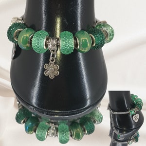 May include: A green beaded bracelet with various textures and shades of green beads. The bracelet includes sparkling, smooth, and patterned beads, along with a silver flower charm. The bracelet is displayed on a black stand.