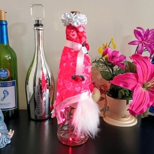 May include: A collection of decorative wine bottles and accessories. Includes a green wine bottle with a blue label, a silver bottle with a clear stopper, and a bottle dressed in red fabric with hearts. A gnome figurine and flowers are also present.