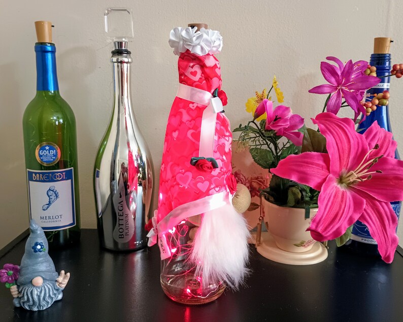 May include: A collection of decorative wine bottles and accessories. Includes a green wine bottle with a blue label, a silver bottle with a clear stopper, and a bottle dressed in red fabric with hearts. A gnome figurine and flowers are also present.