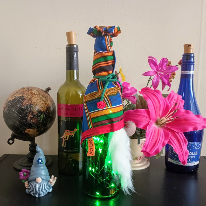May include: A decorative wine bottle with a gnome design. The bottle is wrapped in colorful fabric and has a white beard. The bottle is lit with green fairy lights. A small gnome figurine and a globe are also in the image.