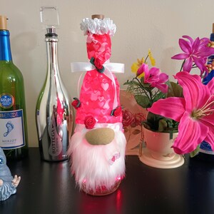 May include: A collection of decorative wine bottles and a gnome figurine. The bottles are green, silver, and blue, with various stoppers. A gnome with a white beard and a red heart-patterned hat is illuminated. Pink flowers and a small gnome figurine are also present.