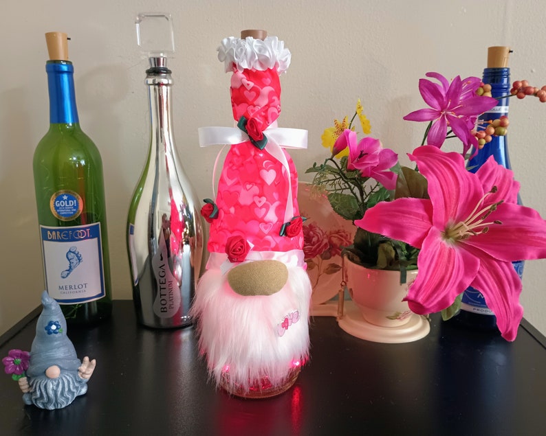 May include: A collection of decorative wine bottles and a gnome figurine. The bottles are green, silver, and blue, with various stoppers. A gnome with a white beard and a red heart-patterned hat is illuminated. Pink flowers and a small gnome figurine are also present.
