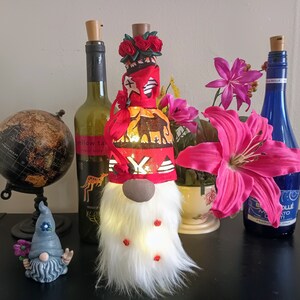 May include: A decorative gnome bottle topper with a white faux fur beard and a red hat with an African safari animal print. The gnome has a brown nose and small red accents. A globe, wine bottles, and flowers are in the background.