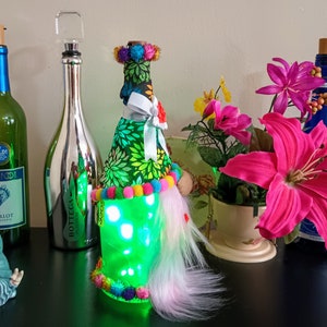 May include: A collection of decorative wine bottles and accessories. Includes a green bottle with a blue label, a silver bottle, and a bottle adorned with fabric, lights, and colorful pom-poms. A small gnome figurine and flowers are also present.