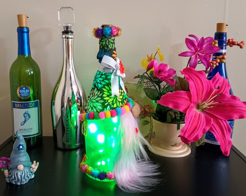 May include: A collection of decorative wine bottles and accessories. Includes a green bottle with a blue label, a silver bottle, and a bottle adorned with fabric, lights, and colorful pom-poms. A small gnome figurine and flowers are also present.