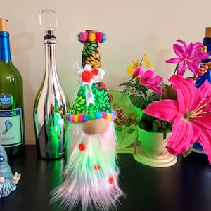 May include: A collection of decorative wine bottles and a gnome figurine. The bottles include a green Barefoot Merlot bottle, a silver Bottega Platinum bottle, and a blue bottle. A colorful gnome with a long beard and a pointed hat is also present.