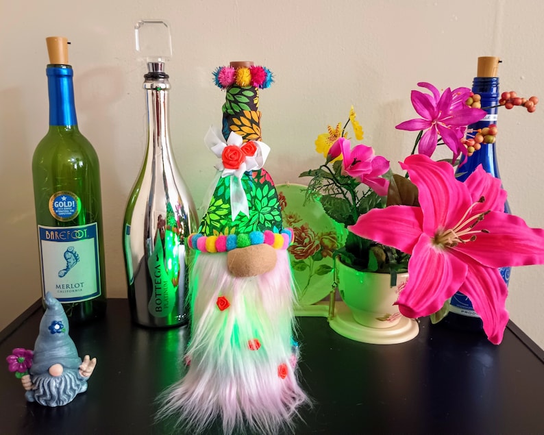 May include: A collection of decorative wine bottles and a gnome figurine. The bottles include a green Barefoot Merlot bottle, a silver Bottega Platinum bottle, and a blue bottle. A colorful gnome with a long beard and a pointed hat is also present.