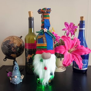 May include: A whimsical wine bottle gnome with a white beard, red hat, and colorful fabric hat. The gnome sits atop a wine bottle with green lights. Other bottles and a small gnome figurine are in the background.