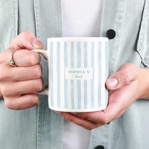 May include: White ceramic mug with vertical light blue stripes. The mug has the text "SOPHIA'S dad" in a rectangular white box. The mug is held by a person wearing a light green jacket.
