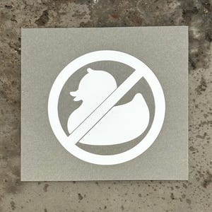 May include: White sticker with a circle and a diagonal line through it. Inside the circle is a white silhouette of a rubber duck.