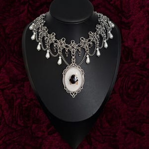 May include: A silver-toned chainmail choker necklace featuring a central oval pendant with a white background and a black insect motif. The necklace is adorned with multiple white teardrop-shaped beads hanging from the silver chain links.