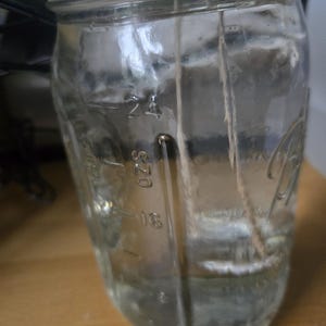 Könnte beinhalten: Ein klares Glasgefäß mit einem weißen Deckel, gefüllt mit einer klaren Flüssigkeit und zwei weißen Schnüren, die herunterhängen. Das Gefäß ist an der Seite mit Markierungen versehen, die 20 und 24 Unzen anzeigen.