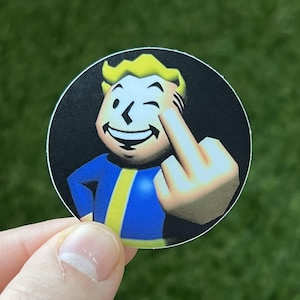 Könnte beinhalten: Runder Aufkleber mit einer Comicfigur in einem blau-gelben Overall, die zwinkert und den Mittelfinger zeigt. Die Figur hat blondes Haar und befindet sich vor einem schwarzen Hintergrund.